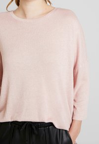 Jasnoróżowy sweter z dzianiny o okrągłym dekolcie i rękawach trzyczwartowych, zestawiony z czarnymi spodniami ze sznurkiem. Miękka faktura, luźny krój.