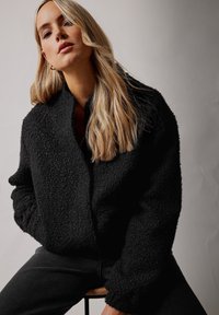 Svart, texturerad långärmad jacka med en bred krage, som har en avslappnad passform och minimalistisk design. Bärs över en svart outfit.