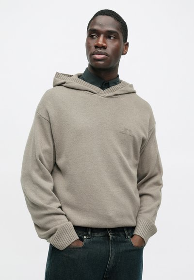 Μπεζ πλεκτή κουκούλα με σχέδιο V-λαιμού, ριγέ μανίκια και διακριτική λεπτομέρεια λογότυπου στο στήθος, φορεμένη πάνω από ένα σκούρο πουκάμισο με γιακά.