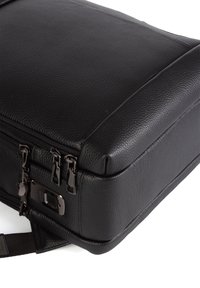 Kulma mustasta tekstiilitaskusta, jossa on kolme metallista vetoketjua ja suorakulmainen lukko yksinkertaisella valkoisella taustalla.