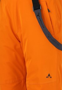 Pantalons d'extérieur orange avec une texture lisse, présentant un logo noir et une sangle réglable grise. Comprend une poche latérale avec une fermeture éclair.