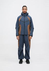 Marineblauwe en bruine weerbestendige jas met capuchon, featuring hoekige ontwerplijnen en ritszakken, gecombineerd met bijpassende broek.