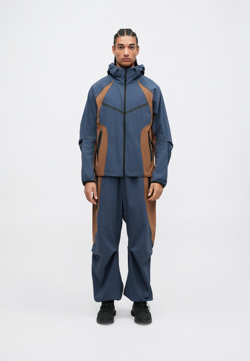 Marineblauwe en bruine weerbestendige jas met capuchon, featuring hoekige ontwerplijnen en ritszakken, gecombineerd met bijpassende broek.