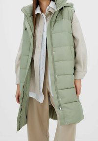 Gewatteerd vest in lichtgroen, met een afneembare capuchon, drukknoopsluiting en een lange pasvorm. Draagt over een beige trui en een wit overhemd.