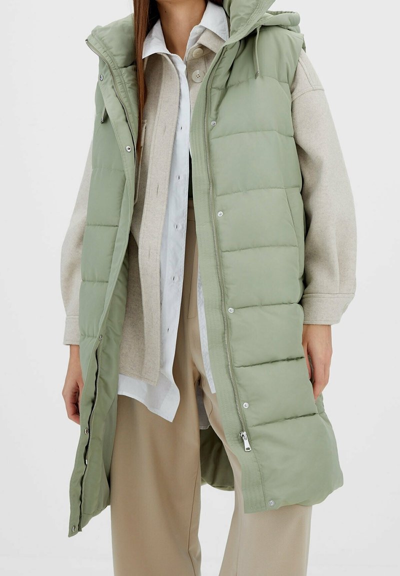 Gewatteerd vest in lichtgroen, met een afneembare capuchon, drukknoopsluiting en een lange pasvorm. Draagt over een beige trui en een wit overhemd.