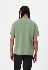 Grön bomull t-shirt med kort ärm och rund halsringning, synlig sömnad och avslappnad passform, sedd bakifrån mot en vit bakgrund.
