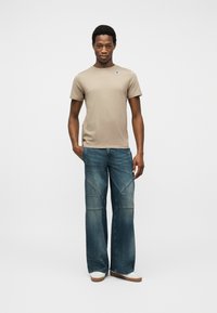 Νεαρός άνδρας που στέκεται, φοράει  μπεζ κοντομάνικη μπλούζα με μικρό λογότυπο, φαρδιά μπλε τζιν και λευκά παπούτσια σε απλό φωτεινό φόντο.