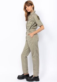 Mono caqui de manga corta, con bolsillos utilitarios y cintura ajustada; presenta una insignia en el hombro izquierdo y piernas ajustadas. Zapatos de plataforma negros.