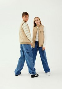 Gilet senza maniche beige sopra camicie a maniche lunghe bianche a righe, abbinati a jeans cargo azzurri larghi. Sneakers in bianco e nero.
