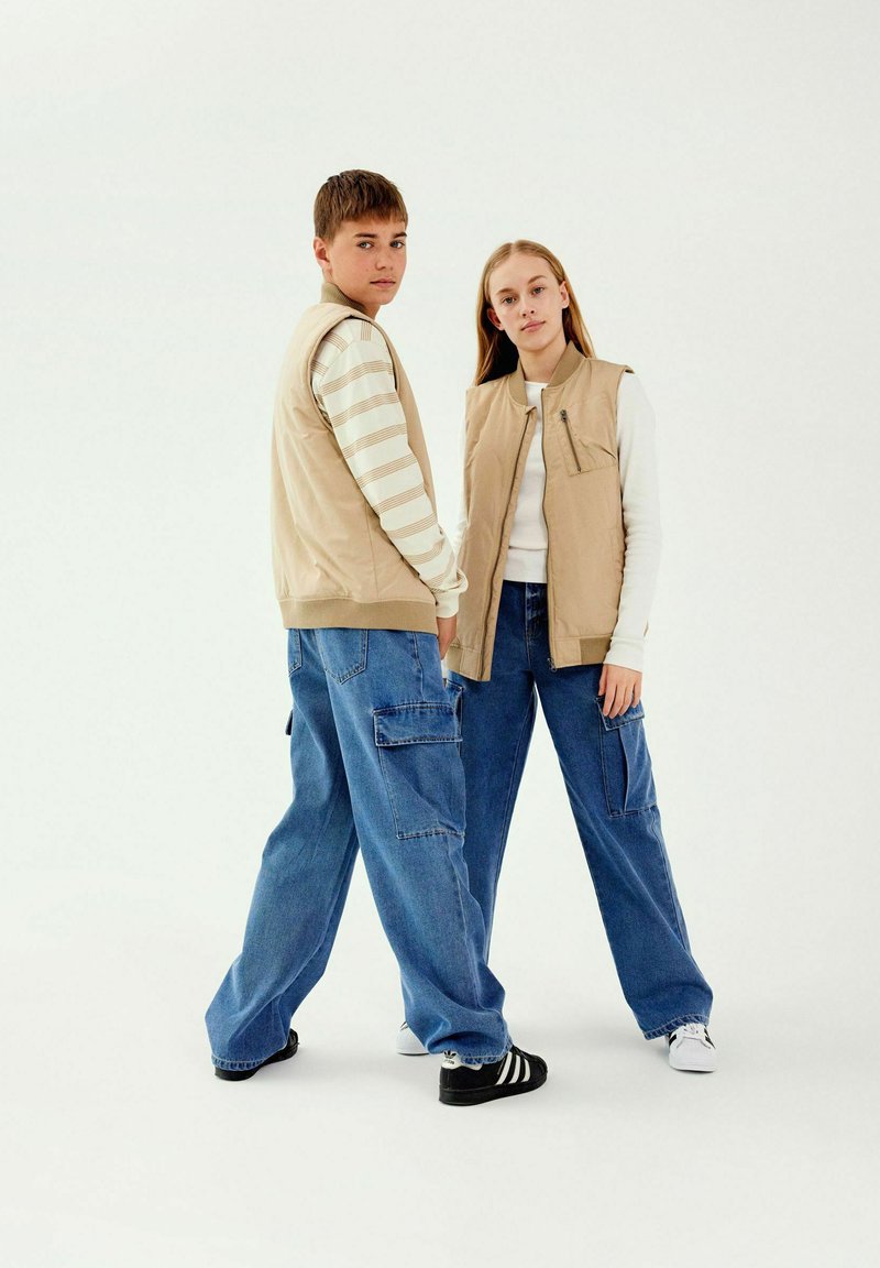 Gilet senza maniche beige sopra camicie a maniche lunghe bianche a righe, abbinati a jeans cargo azzurri larghi. Sneakers in bianco e nero.