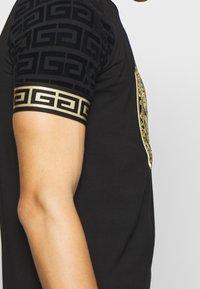 Camisa negra de manga corta con un diseño de logo dorado en el frente. Las mangas tienen un patrón negro texturizado y un acento en dorado.
