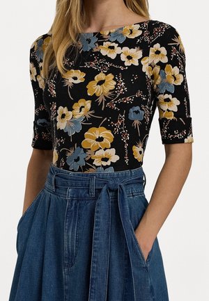 Femme portant un haut noir à fleurs jaunes, bleues et crème et une jupe en denim bleu taille haute avec une ceinture nouée et des poches.