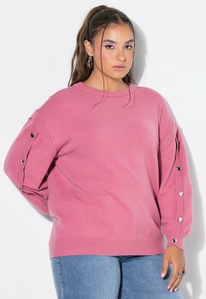 Ung kvinde med langt hår iført en lyserød sweater med hjerteformede sølvknapper på ærmerne og lyseblå jeans mod hvid baggrund.