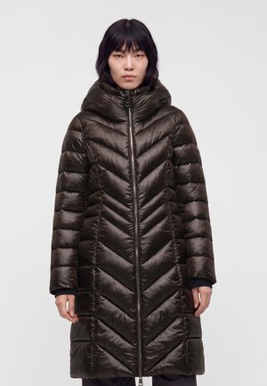 Femme portant un long manteau matelassé marron foncé avec un col montant et une fermeture éclair à l'avant, debout devant un fond clair uni.