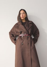Cappotto di lana marrone con chiusura doppiopetto, grandi bottoni neri e cintura bordeaux a contrasto in vita. Tessuto liscio e vestibilità morbida.