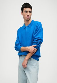 Maglione polo blu con colletto e chiusura a bottoni, in tessuto a maglia con polsini e orlo a coste, abbinato a jeans azzurri chiari.