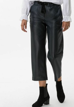 Person iført sorte cropped læderbukser med snøre i taljen, hvid bluse med flæsemanchetter og sorte ankelstøvler med hæl.