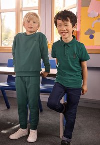 To børn står i et klasseværelse. Den ene har en grøn sweatshirt og matchende joggingbukser på, den anden en grøn polo og mørke jeans. Hvide og sorte sko.