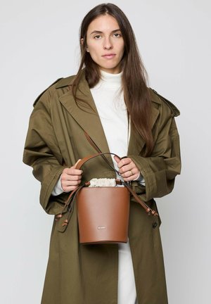 Woman with long brown hair wearing an olive trench coat and white turtleneck, holding a brown leather bucket bag with drawstring top.