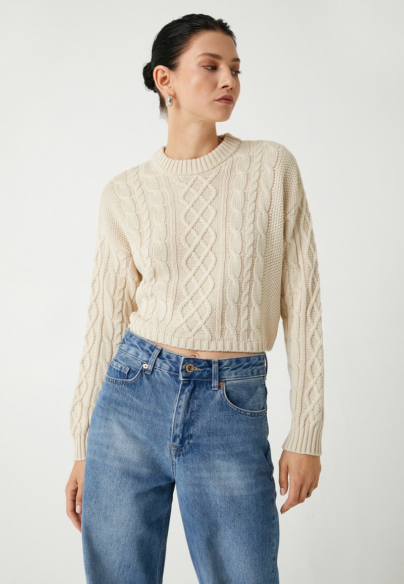 Maglione a trecce color crema con scollo rotondo e orlo a coste, abbinato a jeans in denim blu a vita alta. Dettagli del motivo testurizzato visibili.
