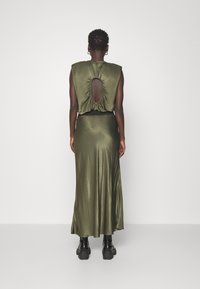 Abito in satin verde oliva con corpino aderente, scollatura a barco e gonna fluida fino alla caviglia, abbinato a stivali con piattaforma neri.