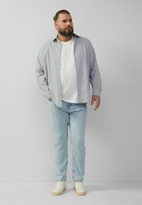 Lichtgrijs overhemd met knopen over een wit T-shirt, gecombineerd met lichtblauwe jeans en off-white sneakers. Model staat met handen in de zakken.