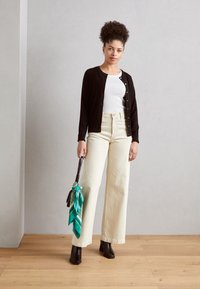 Zwarte cardigan met gouden knopen, witte aansluitende top, crèmekleurige high-waisted wijde broek en donkere enkellaarsjes. Groene gebloemde sjaal op handbagage.