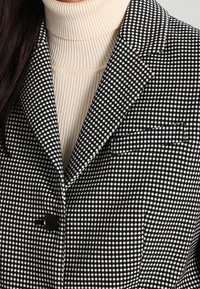 Svart och vit houndstooth-blazer med notch-lapels, enkel svart knappstängning och bröstficka, lager på en ribbad beige polotröja.