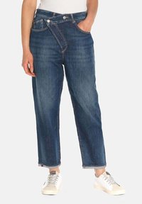 Blauwe spijkerbroek met een rechte pasvorm, hoge taille en unieke kruisknoopsluiting. Draagt met witte sneakers.