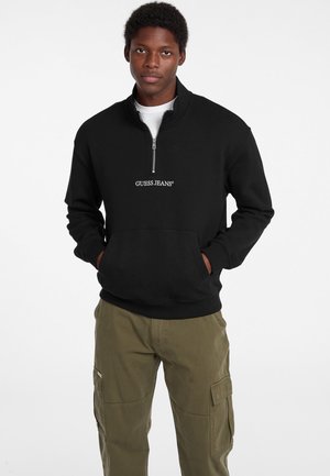 Giovane uomo che indossa una felpa nera Guess Jeans con mezza cerniera e pantaloni cargo verde oliva, in piedi con le mani nelle tasche.