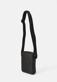 Sac bandoulière noir de forme rectangulaire, en tissu texturé, avec une bandoulière réglable. Il possède une fermeture éclair et un accent logo discret.