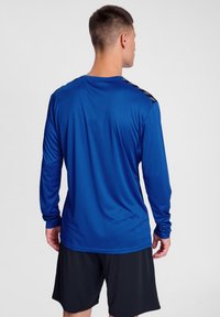 Langärmliges, blaues Sportshirt aus leichtem, glattem Stoff. Besitzt schwarze Akzente an den Schultern und einen lockeren Schnitt. Kombiniert mit schwarzen Shorts.