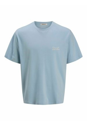 Egy fiú áll és mosolyog, világoskék pólót, farmer rövidnadrágot, fehér zoknit és fekete-fehér sportcipőt visel, sima fehér háttér előtt.