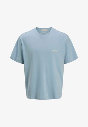 Chłopiec stoi i się uśmiecha, ubrany w jasnoniebieski t-shirt, jeansowe spodenki, białe skarpetki i czarno-białe trampki, na jednolitym białym tle.