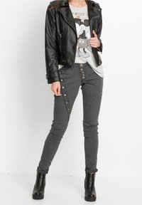 Černá kožená zkrácená bunda přes šedé tričko s potiskem, na kterém je karikatura. Šedé úzké kalhoty s detailem knoflíků na boku. Černé kotníčkové boty.