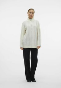 Pulover iz kabelskega pletenja v off-white barvi z visokim ovratnikom in četrtinskim zadrgo, v kombinaciji z črnimi širokimi hlačami in črnimi natikači.
