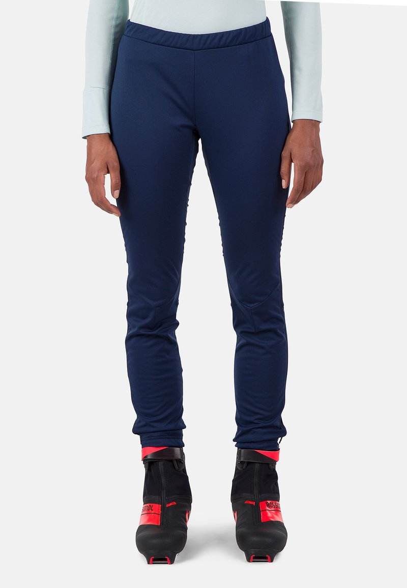Pantaloni sportivi blu navy con vita elastica, realizzati in un tessuto liscio ed elastico, abbinati a stivali da sci neri con dettagli rossi.