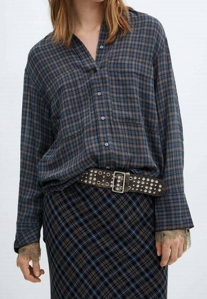 Femme portant une chemise à carreaux bleue et marron boutonnée, rentrée dans une jupe à carreaux foncée avec une ceinture cloutée et des poignets ornés de dentelle.