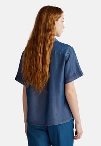 Camisa de denim, de manga corta, color azul claro, corte relajado, costuras visibles, detalle en el yugo trasero, lleva puesta con pantalones de denim azul.