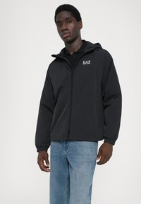 Svart lättviktsjacka med främre dragkedja, huva, elastiska ärmslut och en logotyp på vänster bröst. Bärs över en svart polo och blåjeans.