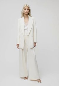 Weißes maßgeschneidertes Blazer über einem weißen Satinoberteil mit Spitze, kombiniert mit weiten Hosen. Glatter Stoff, klare Linien, minimales Design.