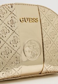 Guldfarvet Guess clutch med præget logo mønster, glat tekstur og cirkulær detalje. Lynlåslukning med detaljeret syning.