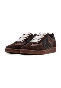 Sneaker in suede marrone con accenti in tessuto nero, lacci bianchi e suola in gomma. Presenta un elemento di design laterale e un sottile dettaglio di branding.