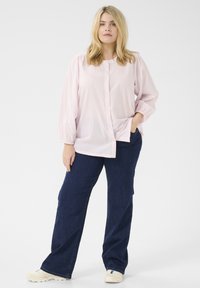 Camicia button-up rosa chiaro con texture a pois, maniche lunghe a sbuffo e due tasche laterali, abbinata a jeans a gamba dritta blu scuro.