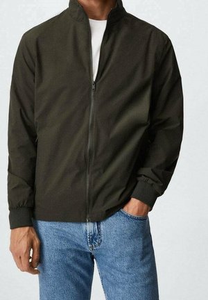 Donkergroene lichtgewicht zip-up jack met een staande kraag, elastische manchetten en een gladde textuur, gecombineerd met lichtblauwe jeans.