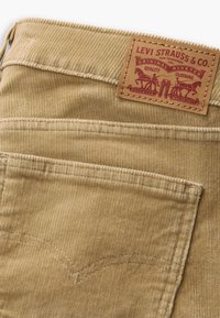 Tan kordfløyelsbukser med et vevd mønster, med et lærmerke preget med "Levi Strauss & Co." og dekorativ søm på lommene.