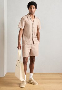 Conjunto de camiseta de manga corta y pantalones cortos en marrón claro con un patrón sutil; combinado con zapatillas beige y un bolso de tela crema. Fondo de suelo de madera.