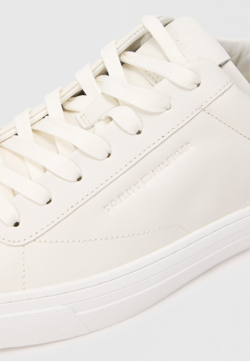 White leather sneaker with round toe, textured upper, flat laces, and embossed "TOMMY HILFIGER" logo on the side. Rubber sole, minimal design.