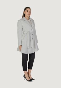 Cappotto grigio chiaro con una trama testurizzata, design doppiopetto, grandi bottoni e vita con cintura. Abbinato a pantaloni neri a vita alta e tacchi.