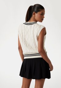 White sleeveless knit sweater with a cable pattern and black stripes around the collar and waistband, paired with a black pleated skirt.